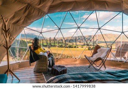 Woman looking out at nature from geo dome tents. Green, blue, orange background. Cozy, camping, glamping, holiday, vacation lifestyle concept. Outdoors cabin, scenic background. New Zealand.