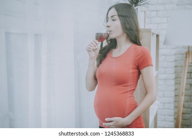 Woman is Looking out from Window. Woman is a Young Brunette Pregnant Girl. Girl is Drinking a Red Wine. Red Wine in Glass. Woman is Touching Her Belly. Person Located at Home Interior.
