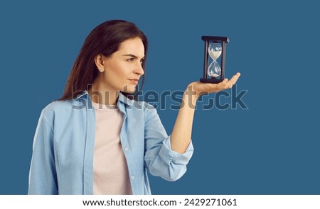 Woman looking at sand glass in her hand. Side view of beautiful young girl in casual shirt standing on dark blue studio background, holding hour glass, looking at falling sand, and thinking about time