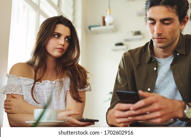 Woman looking unhappy while her man paying no attention to her and busy using his mobile phone. Sulking woman sitting next to man reading text messages during a date. - Powered by Shutterstock