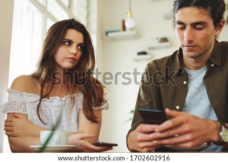 Woman looking unhappy while her man paying no attention to her and busy using his mobile phone. Sulking woman sitting next to man reading text messages during a date.