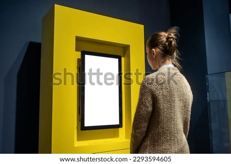 Woman looking at vertical blank digital interactive white display wall at exhibition or museum with futuristic interior. Mock up, future, copyspace, white screen, technology concept