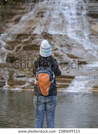woman looking at a waterfall on a nature hike (photographed from behind) wearing winter hat, brown jacket, backpack, blue jeans (buttermilk falls state park in ithaca new york, autumn, bright foliage)