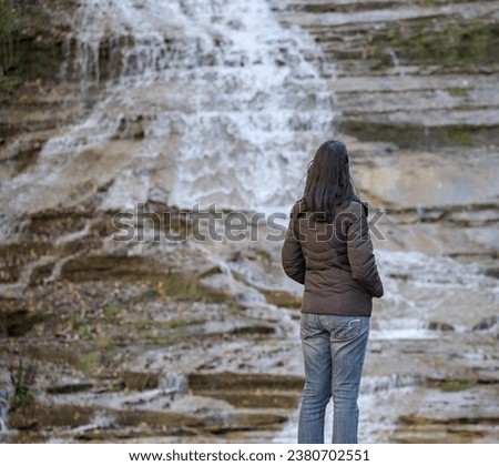 woman looking at a waterfall on a nature hike (photographed from behind) wearing winter hat, brown jacket, blue jeans (buttermilk falls state park in ithaca new york, autumn, bright foliage, gorge)