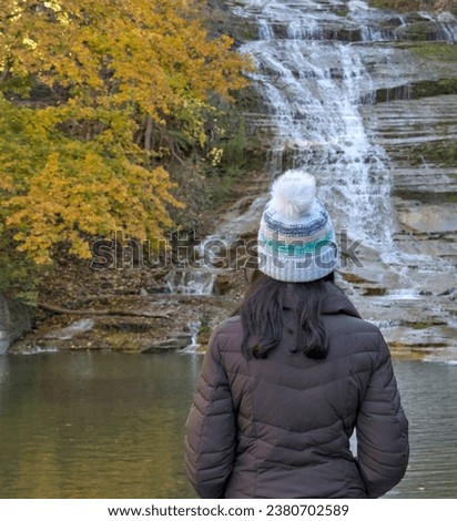 woman looking at a waterfall on a nature hike (photographed from behind) wearing winter hat, brown jacket (buttermilk falls state park in ithaca new york, autumn, leaves changing colors, gorge)