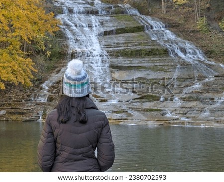 woman looking at a waterfall on a nature hike (photographed from behind) wearing winter hat, brown jacket (buttermilk falls state park in ithaca new york, autumn, bright foliage, gorge trail)