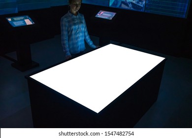 Woman Looking At White Blank Interactive Touchscreen Display Of Electronic Multimedia Table Kiosk In Dark Room Of Technology Museum. Mock Up, Copyspace, Template And Futuristic Concept