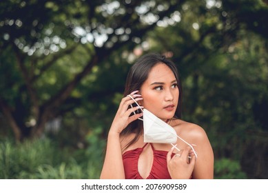 A Woman Looks Around Nervously While Taking Off Her Face Mask. A Bit Hesitant And Unaccustomed Going Mask-less. Outdoor Scene.