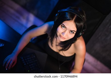 A woman looks at camera while sitting in front of her PC and playing online computer games and streaming game on the internet. Gamer girl plays at the table in his room. Top view.