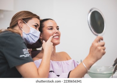 The woman looks at her smile with a mirror in a clinic. Dental clinic. Well-groomed white teeth after being treated by a doctor. the woman dentist points to the smile of the patient. - Powered by Shutterstock