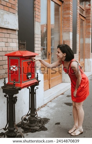 A woman looks into a red street mailbox. The girl throws a letter into an old mailbox. Paper letters correspondence. Send a letter with interesting facts about your trip through the mailbox.