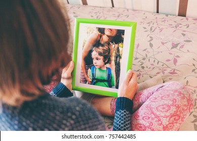 A Woman Looks At A Photo Of A Boy. Mom Holds A Photo Frame With A Photograph Of Her Son. A Small Child And Memories.