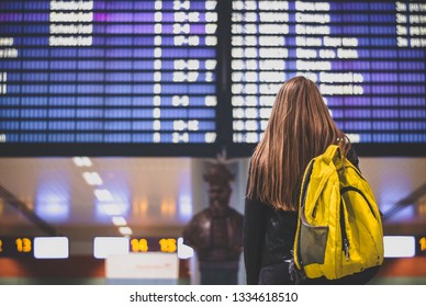 A Woman Looks At The Screen With A Schedule. Passenger With Baggage At The Airport. Back View.