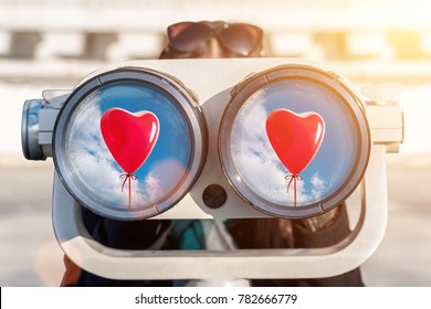 A Woman Looks Through Binoculars. In The Reflection Of The Glass You Can See Red Heart Balloon. Sunny.