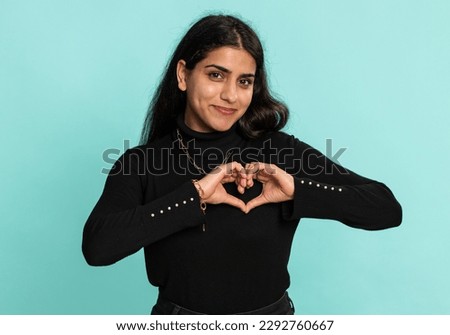 Woman in love. Smiling arabian indian woman 20 years old makes heart gesture demonstrates love sign expresses good feelings and sympathy. Pretty hindu girl isolated alone on blue studio background