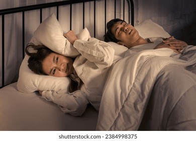 woman lying awake in bed, covering her ears to block out the sound of her partner snoring, capturing a common sleep disturbance