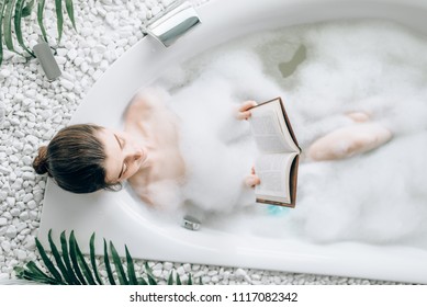 Woman lying in bath with foam and reads magazine