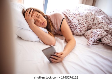 Woman Lying In Bed Reaching To Turn Off The Alarm On Her Mobile Phone In Morning. Sleepy Woman Being Woken Up By Smartphone Alarm. Young Woman Waking Up In Bed And Checking Her Smartphone