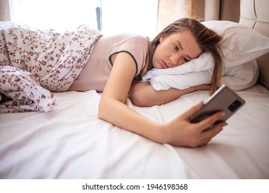 Woman Lying In Bed Reaching To Turn Off The Alarm On Her Mobile Phone In Morning. Sleepy Woman Being Woken Up By Smartphone Alarm. Young Woman Waking Up In Bed And Checking Her Smartphone