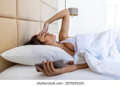 Woman Lying In Bed Reaching To Turn Off The Alarm On Her Mobile Phone In Morning. Sleepy Woman Being Woken Up By Smartphone Alarm. Young Woman Waking Up In Bed And Checking Her Smartphone