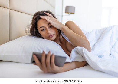 Woman Lying In Bed Reaching To Turn Off The Alarm On Her Mobile Phone In Morning. Sleepy Woman Being Woken Up By Smartphone Alarm. Young Woman Waking Up In Bed And Checking Her Smartphone