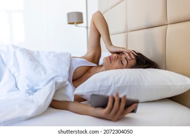 Woman Lying In Bed Reaching To Turn Off The Alarm On Her Mobile Phone In Morning. Sleepy Woman Being Woken Up By Smartphone Alarm. Young Woman Waking Up In Bed And Checking Her Smartphone