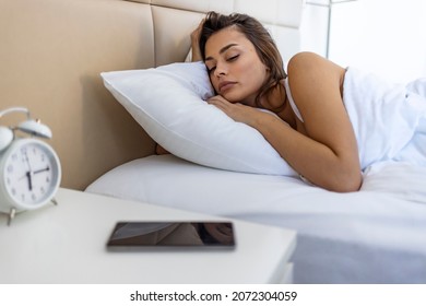 Woman Lying In Bed Reaching To Turn Off The Alarm On Her Mobile Phone In Morning. Sleepy Woman Being Woken Up By Smartphone Alarm. Young Woman Waking Up In Bed And Checking Her Smartphone