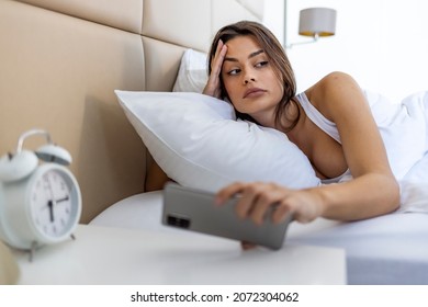 Woman Lying In Bed Reaching To Turn Off The Alarm On Her Mobile Phone In Morning. Sleepy Woman Being Woken Up By Smartphone Alarm. Young Woman Waking Up In Bed And Checking Her Smartphone