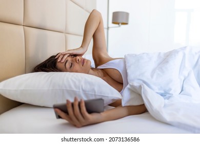 Woman Lying In Bed Reaching To Turn Off The Alarm On Her Mobile Phone In Morning. Sleepy Woman Being Woken Up By Smartphone Alarm. Young Woman Waking Up In Bed And Checking Her Smartphone