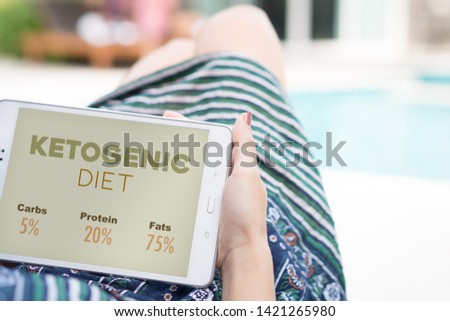 A woman lying down relaxing on a day bed beside the swimming pool reading the information about Ketogenic Diet on tablet computer. Online Application, Digital device, Keto Diet info. 
