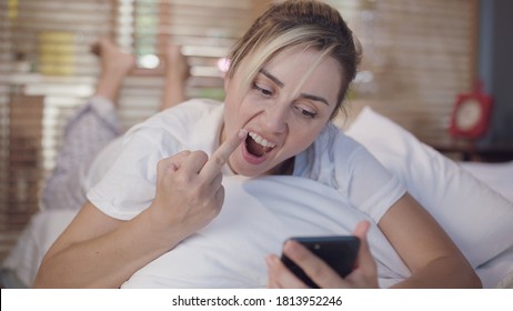 The woman lying face down on her bed in white sheets on the phone gets angry at the news she sees on her phone.Middle finger, rude sign of disrespect, woman making a damn gesture with angry look.