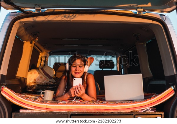 Woman lying face down on the bed of her camper van\
while using mobile to listen to music. Front shot of a young girl\
enjoying the sunset with her cup of tea or coffee and her laptop.\
Digital nomad