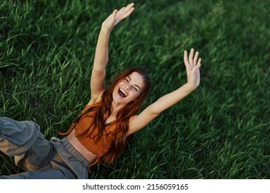 A woman lying in nature on the green grass in the park resting with her hands up and smiling with her mouth open, her red hair flying in the wind in the open air in the sunlight. Freelancer's concept - Powered by Shutterstock