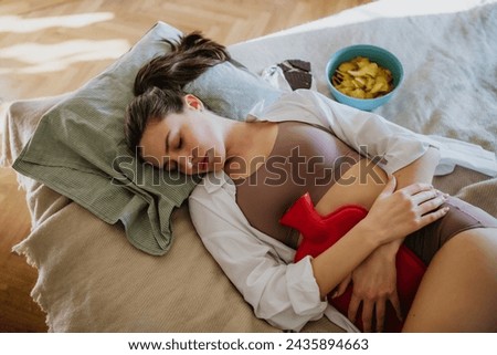 Woman lying on bed with menstrual pain and cramps, having period cravings. Woman warming lower abdomen with a hot water bottle, endometriosis, and conditions causing pain in tummy.