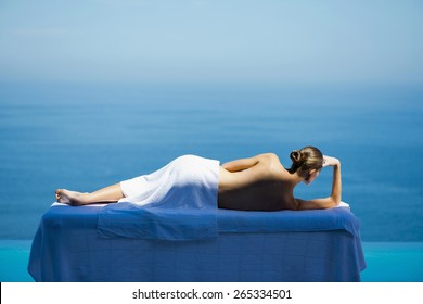 Woman lying on blue massage table facing sea. Rear view.