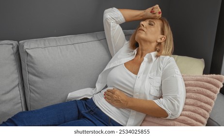 Woman lying on a couch holding her stomach with a pained expression in a serene living room setting wearing casual clothes - Powered by Shutterstock