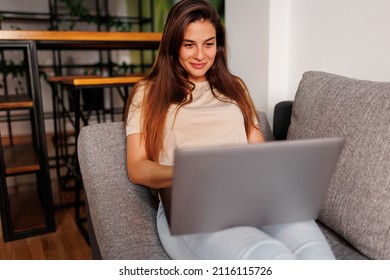 Woman lying on the couch with laptop in her lap, relaxing at home and chatting online with friends
