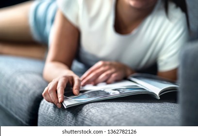 Woman Lying On Couch And Reading A Fashion Or Beauty Magazine. Lady Enjoying Her Day Off With Celebrity Entertainment News Or Shopping Catalogue. Hipster Girl Relaxing On Sunday Morning On Comfy Sofa.