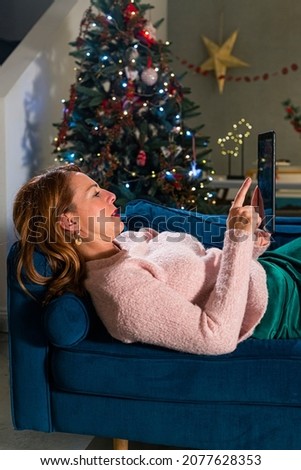 Woman is lying on the couch with a tablet in her hands, while a pet dog is next to her. The living room is decorated in the spirit of the Christmas holidays.