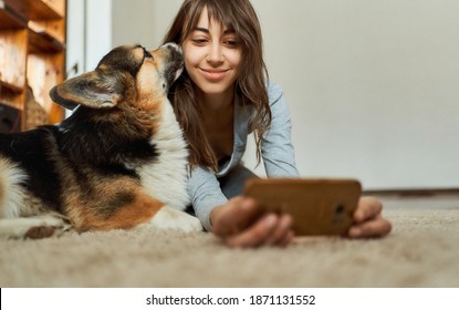 Woman Lying On Floor In Living Room With Pet And Making Selfie Photo, Welsh Corgi Dog Kissing His Owner