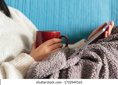 Woman lying on sofa with blanket watching a movie on tablet and holding a mug