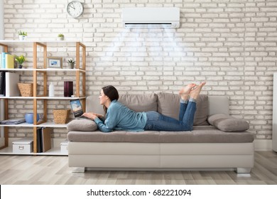 Woman Lying On Sofa Using Laptop Enjoying The Cooling Of Air Conditioner At Home