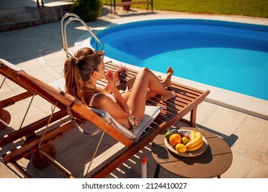 Woman lying on sunbed by the swimming pool, eating grapes, sunbathing and relaxing on summer vacation