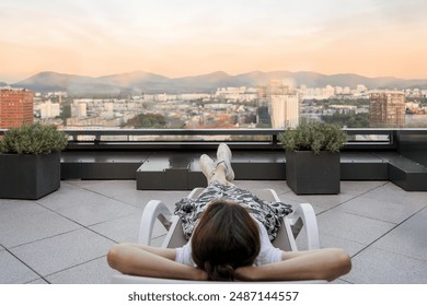 Woman lying on sunbed on spacious summer terrace with view on mountains and cityscape. Rooftop relaxing zone with potted plants. Private area in hotel - Powered by Shutterstock