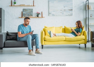 woman lying on yellow sofa with smartphone near husband sitting on couch and using smartphone - Shutterstock ID 1491876143