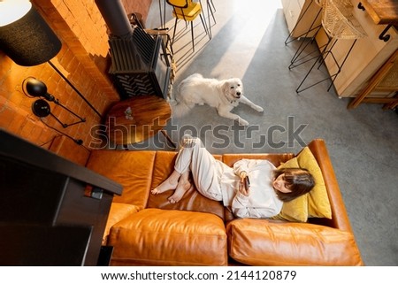 Woman lying relaxed on couch and using phone with her dog at home. Wide angle view from above. Spending leisure time with digital gadgets and pet