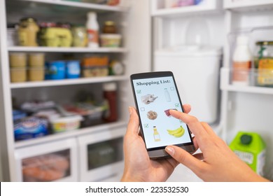 Woman makes her shopping list on his phone connected to the refrigerator