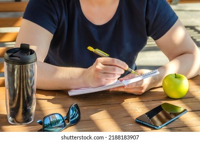 Woman Makes Notes In Notebook During Lunch, Sitting On Veranda On Sunny Day. Apple And Coffee In Tumbler On Table