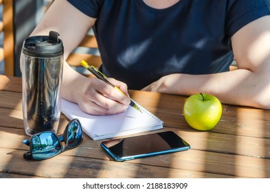 Woman Makes Notes In Notebook During Lunch, Sitting On Veranda On Sunny Day. Apple And Coffee In Tumbler On Table