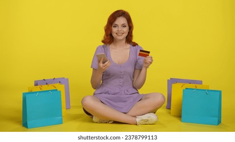 A woman makes a purchase in an online store using a smartphone and a credit card. Redhaired woman sits crosslegged on the floor surrounded by shopping bags on a yellow background.